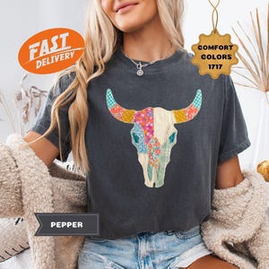 May include: A charcoal gray t-shirt featuring a colorful floral and patchwork design of a bull skull. The shirt has short sleeves and a crew neck. The text "Pepper" is on a banner. The image also includes a "Fast Delivery" and "Comfort Colors 1717" tag.