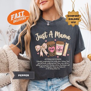 May include: A dark gray t-shirt with the text "Just A Mama" and coffee cup graphics. The shirt features a bow, hearts, and the phrase "I love coffee." Additional text includes "#STRINGCHEESEMAMA" and a list of words. The shirt is part of the Comfort Colors collection.