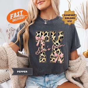 May include: Dark grey t-shirt featuring the letters "TX" in a leopard print pattern, embellished with pink bows and the cursive text "She's from". The shirt is part of the "Comfort Colors 1717" collection.