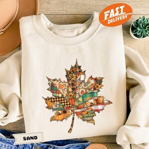 Op de afbeelding: Crèmekleurige sweatshirt met een groot, kleurrijk bladontwerp gemaakt van patchworkpatronen. Het blad bevat herfstthema-elementen zoals pompoenen en zonnebloemen. Het woord "SAND" staat op een klein label.