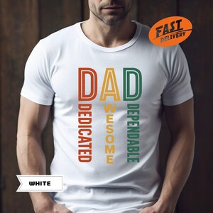 K&ouml;nnte beinhalten: Wei&szlig;es T-Shirt mit dem Wort "DAD" in Orange, "AWESOME" in Gelb und "DEPENDABLE" in Gr&uuml;n. Die W&ouml;rter "DEDICATED" sind vertikal in Orange ausgerichtet. Das Shirt hat auch einen "FAST DELIVERY" Aufkleber.