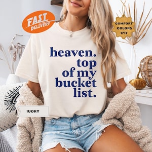 以下が含まれることがあります： 「heaven. top of my bucket list.」と紺色の文字がプリントされたアイボリー色のTシャツ。Comfort Colorsコレクションの一部です。モデルはデニムショーツとクリーム色のカーディガンを着用しています。