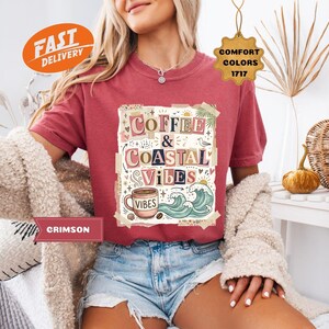 May include: Crimson-colored t-shirt with the words "Coffee & Coastal Vibes" in a decorative design. The shirt features a coffee cup, waves, and palm leaves. The shirt is paired with denim shorts.