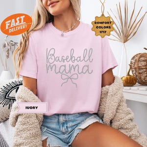 May include: A light pink t-shirt with the text "Baseball Mama" in a dotted design. The shirt has short sleeves and is paired with denim shorts. A cream-colored sweater is draped over the shoulders.