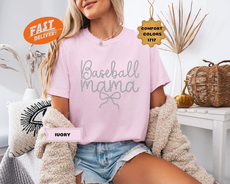 May include: A light pink t-shirt with the text "Baseball Mama" in a dotted design. The shirt has short sleeves and is paired with denim shorts. A cream-colored sweater is draped over the shoulders.