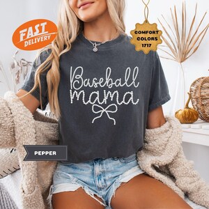 May include: Dark gray t-shirt with the text "Baseball Mama" in a white, beaded font. The shirt has a bow design under the text. The model is wearing denim shorts and a cream-colored cardigan. The shirt is part of the Comfort Colors 1717 collection.