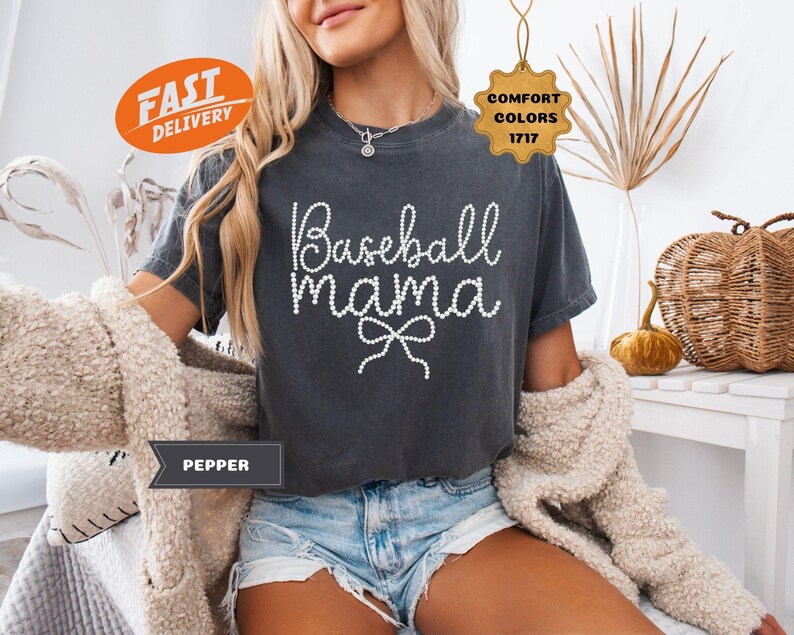 May include: Dark gray t-shirt with the text "Baseball Mama" in a white, beaded font. The shirt has a bow design under the text. The model is wearing denim shorts and a cream-colored cardigan. The shirt is part of the Comfort Colors 1717 collection.