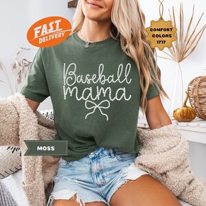 May include: Moss green t-shirt with the text "Baseball Mama" in white script, accented with a bow. The shirt is part of the Comfort Colors 1717 collection. The model is wearing denim shorts and a beige cardigan.