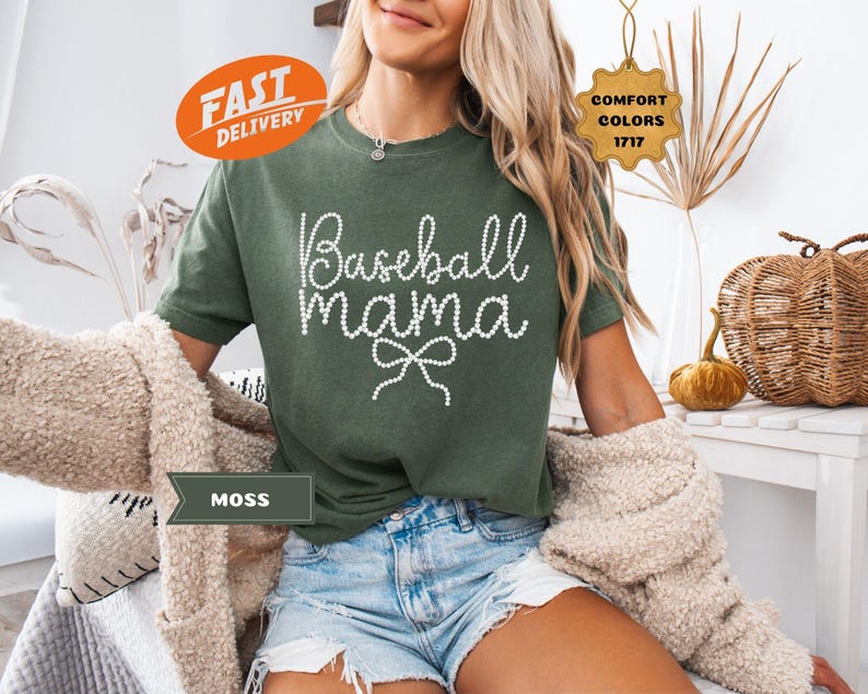 May include: Moss green t-shirt with the text "Baseball Mama" in white script, accented with a bow. The shirt is part of the Comfort Colors 1717 collection. The model is wearing denim shorts and a beige cardigan.