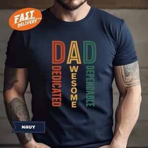 K&ouml;nnte beinhalten: Marineblaues T-Shirt mit dem Wort "DAD" in Orange, "AWESOME" in Gelb und "DEPENDABLE" in Gr&uuml;n. Das Wort "DEDICATED" ist vertikal in Orange gedruckt. Das Shirt hat einen "FAST DELIVERY" Aufkleber.