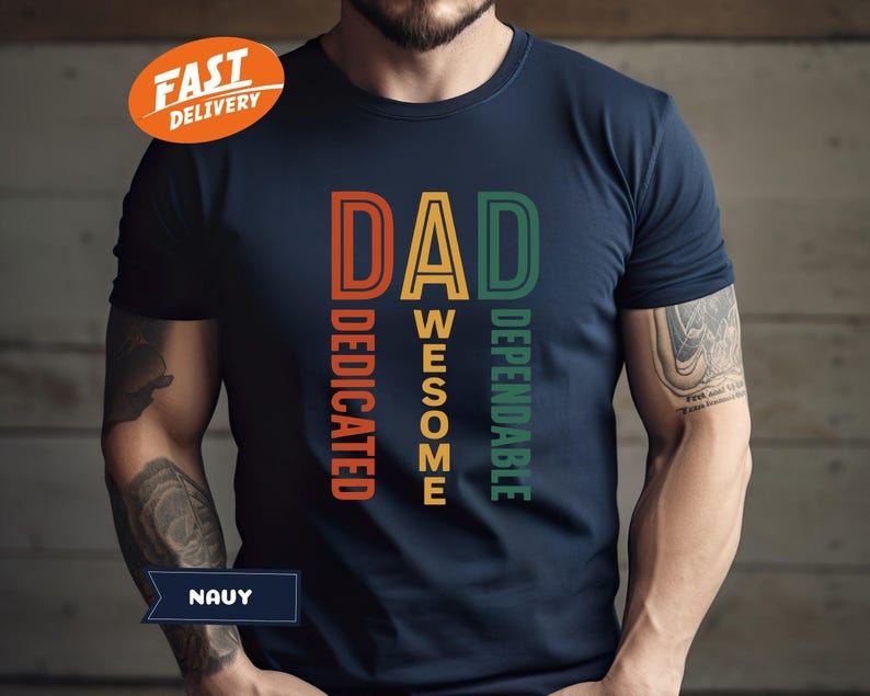 K&ouml;nnte beinhalten: Marineblaues T-Shirt mit dem Wort "DAD" in Orange, "AWESOME" in Gelb und "DEPENDABLE" in Gr&uuml;n. Das Wort "DEDICATED" ist vertikal in Orange gedruckt. Das Shirt hat einen "FAST DELIVERY" Aufkleber.
