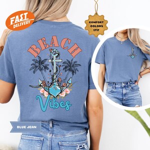 K&ouml;nnte beinhalten: Ein blaues Jeans-T-Shirt mit Strandmotiv. Auf der R&uuml;ckseite des Shirts steht das Wort "BEACH" in korallenfarbenen Buchstaben &uuml;ber einem t&uuml;rkisfarbenen Ankerdesign mit Palmen und dem Wort "Vibes". Auf der Vorderseite befindet sich ein kleinerer Anker.