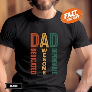 K&ouml;nnte beinhalten: Schwarzes T-Shirt mit dem Wort "DAD" in Orange, "AWESOME" in Gelb und "DEPENDABLE" in Gr&uuml;n. Das Wort "DEDICATED" ist vertikal in Orange gedruckt. Ein "FAST DELIVERY" Aufkleber befindet sich in der oberen rechten Ecke.