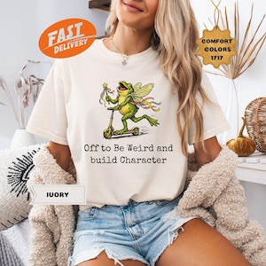 May include: An ivory t-shirt featuring a graphic of a green frog with fairy wings riding a scooter and holding a star wand. Below the frog, the text reads, Off to Be WEIRD and build Character. The shirt has a crew neckline and short sleeves.
