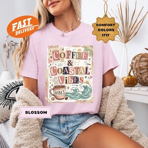 May include: A light pink t-shirt with the words "Coffee & Coastal Vibes" in a colorful, whimsical design. The graphic includes a coffee cup, waves, and other coastal elements. The shirt also has a "Fast Delivery" and "Comfort Colors" tag.