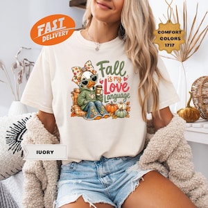 May include: An ivory t-shirt featuring a graphic of a duck wearing a floral headband, sunglasses, and a green hoodie, holding a coffee cup. The text reads Fall is my Love Language, surrounded by pumpkins and autumn leaves. The shirt is labeled as Ivory.