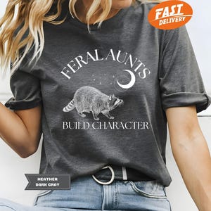 Könnte beinhalten: Ein dunkel-heidesgraues T-Shirt mit einer weißen Grafik eines Waschbären und dem Text "Feral Aunts Build Character".