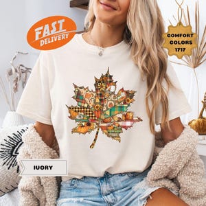 Puede incluir: Camiseta de color marfil con un diseño de hoja de arce grande y colorida. La hoja está compuesta por varios patrones con temática otoñal, incluyendo calabazas, girasoles y cuadros. La camiseta también tiene el texto "FAST DELIVERY", "COMFORT COLORS 1717" y "IUORY".