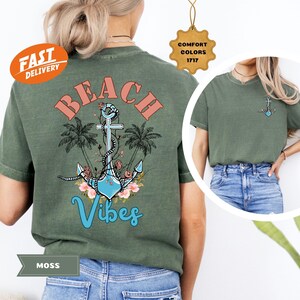 K&ouml;nnte beinhalten: Moosgr&uuml;nes T-Shirt mit Strandmotiv. Das Design zeigt das Wort "BEACH" in korallenfarbenen Buchstaben &uuml;ber einem blauen Anker mit floralen Akzenten und dem Wort "Vibes" in einer Schreibschrift. Palmen umrahmen den Anker.