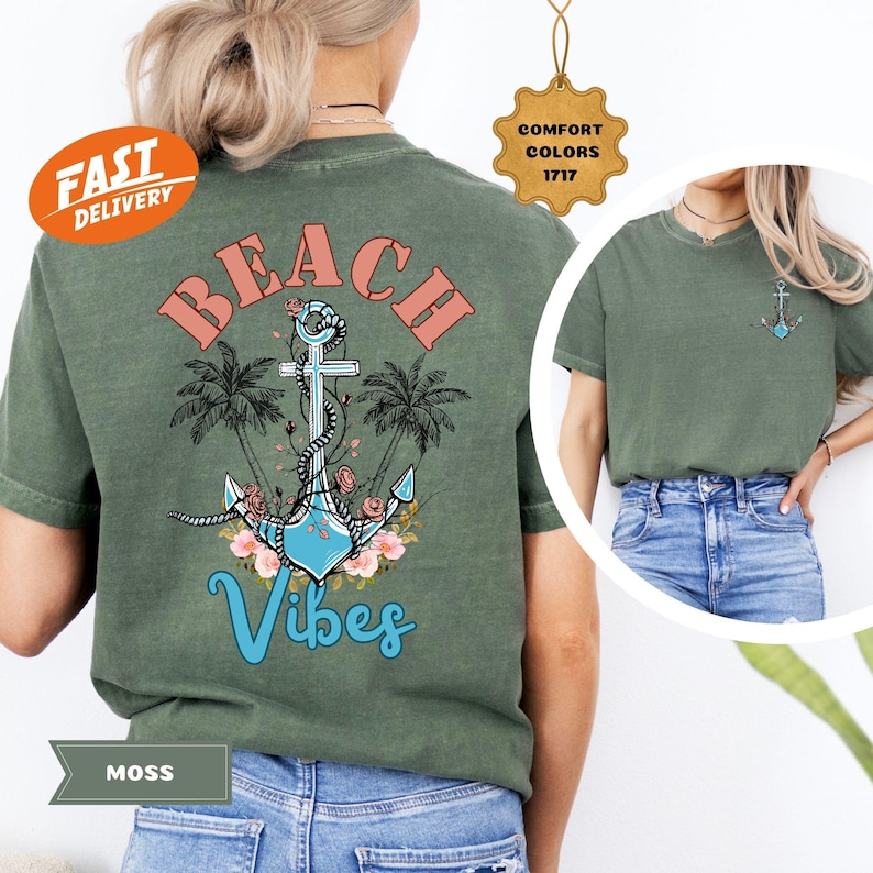 K&ouml;nnte beinhalten: Moosgr&uuml;nes T-Shirt mit Strandmotiv. Das Design zeigt das Wort "BEACH" in korallenfarbenen Buchstaben &uuml;ber einem blauen Anker mit floralen Akzenten und dem Wort "Vibes" in einer Schreibschrift. Palmen umrahmen den Anker.