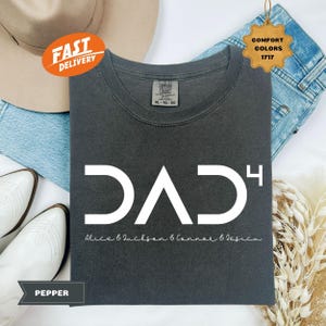May include: A dark gray t-shirt with a white graphic design that says "DAD" with a superscript "4" and the text "Alice & Jackson & Connor & Jessica" below. The shirt is laid flat on a surface with a pair of white cowboy boots and a denim jacket.