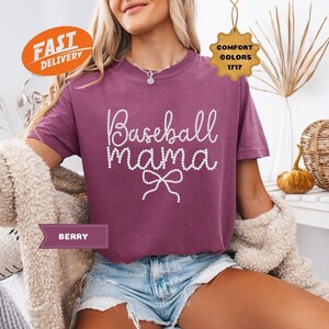 May include: Berry-colored t-shirt with the text "Baseball Mama" in white, embellished with a bow. The shirt is a Comfort Colors brand. The model is wearing denim shorts and a cream-colored cardigan.