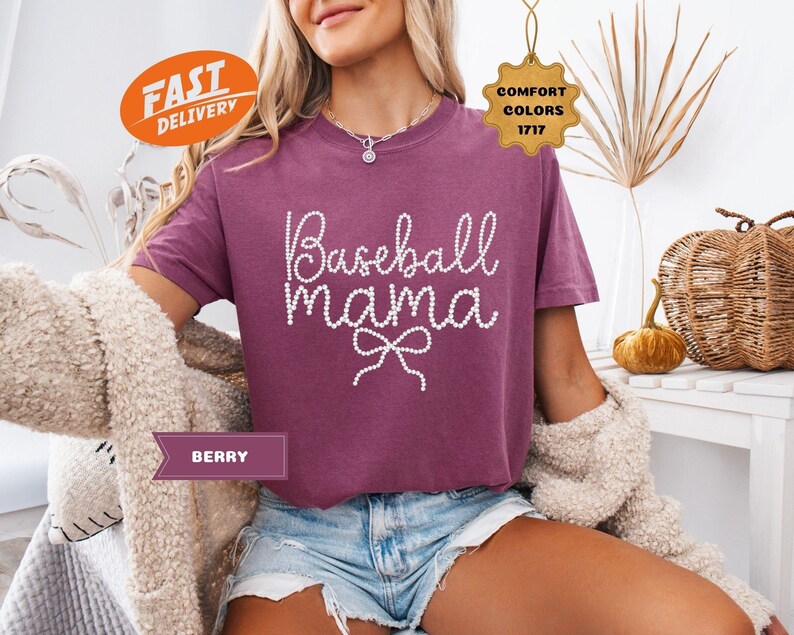 May include: Berry-colored t-shirt with the text "Baseball Mama" in white, embellished with a bow. The shirt is a Comfort Colors brand. The model is wearing denim shorts and a cream-colored cardigan.