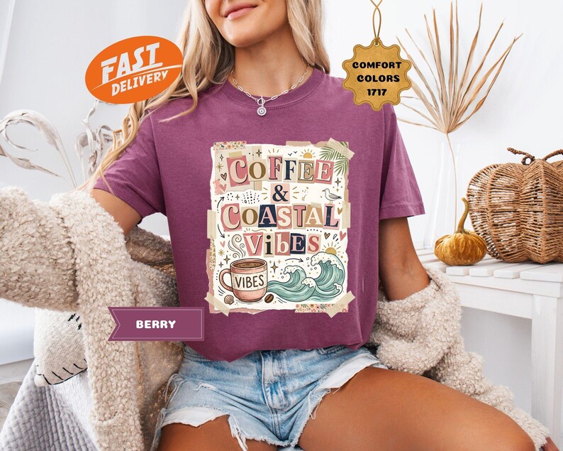 May include: Berry-colored t-shirt with the words "Coffee & Coastal Vibes" in a collage design. The graphic includes a coffee cup, waves, and palm trees. The shirt has a relaxed fit and is made of a soft material.
