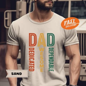 K&ouml;nnte beinhalten: Sandfarbenes T-Shirt mit dem Wort "DAD" in einem vertikalen Design, mit den W&ouml;rtern "DEDICATED", "AWESOME" und "DEPENDABLE" in einem Retro-Farbschema. Ein orangefarbener "FAST DELIVERY" Aufkleber ist sichtbar.