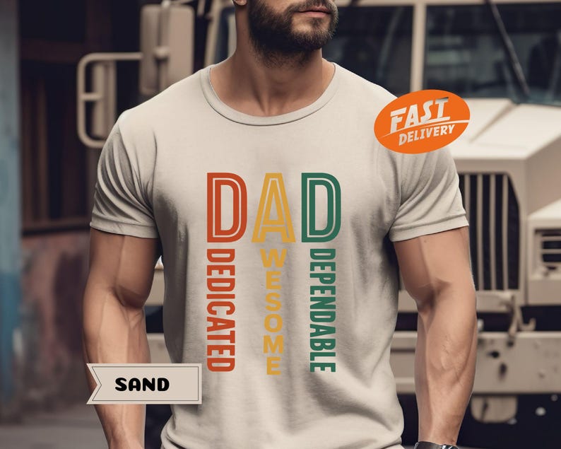 K&ouml;nnte beinhalten: Sandfarbenes T-Shirt mit dem Wort "DAD" in einem vertikalen Design, mit den W&ouml;rtern "DEDICATED", "AWESOME" und "DEPENDABLE" in einem Retro-Farbschema. Ein orangefarbener "FAST DELIVERY" Aufkleber ist sichtbar.