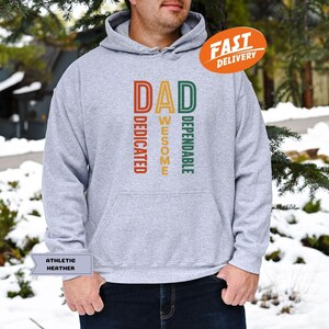 K&ouml;nnte beinhalten: Grauer Kapuzenpullover mit dem Wort "DAD" in Orange, "AWESOME" in Gelb und "DEPENDABLE" in Gr&uuml;n. Das Wort "DEDICATED" ist in Orange. Der Hoodie hat eine Kapuze mit Kordelzug und eine K&auml;ngurutasche. Ein "FAST DELIVERY" Aufkleber ist sichtbar.