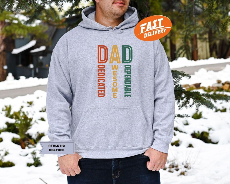 K&ouml;nnte beinhalten: Grauer Kapuzenpullover mit dem Wort "DAD" in Orange, "AWESOME" in Gelb und "DEPENDABLE" in Gr&uuml;n. Das Wort "DEDICATED" ist in Orange. Der Hoodie hat eine Kapuze mit Kordelzug und eine K&auml;ngurutasche. Ein "FAST DELIVERY" Aufkleber ist sichtbar.