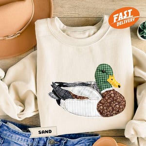 May include: Cream-colored sweatshirt featuring an appliqued duck design. The duck has a green plaid head, a yellow beak, and a brown floral patterned body. The word "SAND" is on a small tag. A brown hat and blue jeans are also in the image.