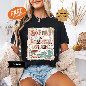 May include: Black t-shirt with the words "Coffee & Coastal Vibes" in a colorful, collage-style design. The graphic includes a coffee cup, waves, and other coastal elements. The shirt is a Comfort Colors 1717.