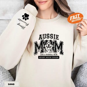 May include: A cream-colored sweatshirt with a black graphic that says "Aussie Mom Like a Normal Mom Except Much Cooler" and a black paw print on the sleeve. The text "Hannah & Princess" is written on the sleeve.