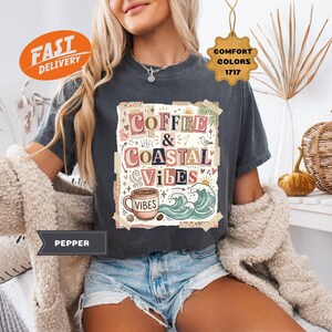 May include: Dark gray t-shirt with the words "Coffee & Coastal Vibes" in a colorful, artistic design. The graphic includes a coffee cup, waves, and palm leaves. The shirt is a comfort color.