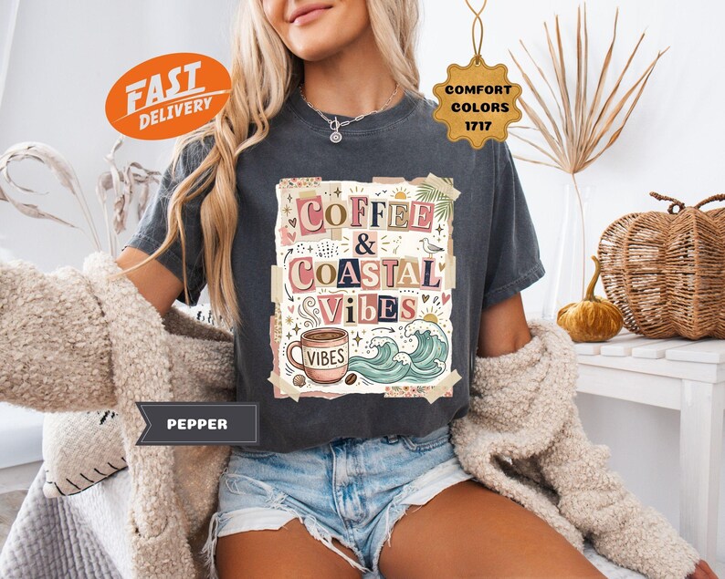 May include: Dark gray t-shirt with the words "Coffee & Coastal Vibes" in a colorful, artistic design. The graphic includes a coffee cup, waves, and palm leaves. The shirt is a comfort color.
