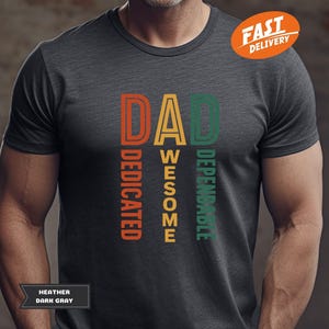 K&ouml;nnte beinhalten: Dunkelgraues T-Shirt mit dem Wort "DAD" in Orange, Gelb und Gr&uuml;n, mit den vertikal aufgedruckten W&ouml;rtern "DEDICATED", "AWESOME" und "DEPENDABLE". Das Shirt tr&auml;gt auch den Text "HEATHER DARK GRAY".