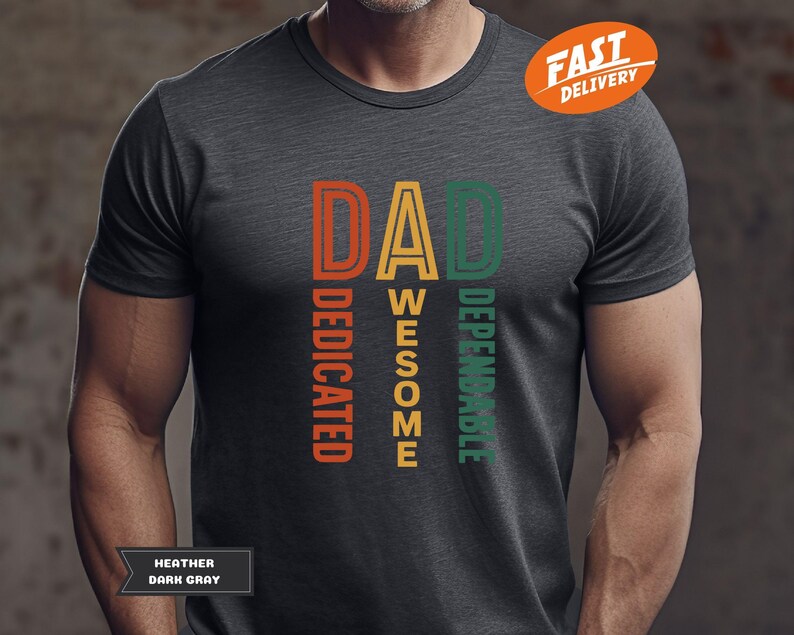 K&ouml;nnte beinhalten: Dunkelgraues T-Shirt mit dem Wort "DAD" in Orange, Gelb und Gr&uuml;n, mit den vertikal aufgedruckten W&ouml;rtern "DEDICATED", "AWESOME" und "DEPENDABLE". Das Shirt tr&auml;gt auch den Text "HEATHER DARK GRAY".