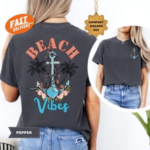 K&ouml;nnte beinhalten: Dunkelgraues T-Shirt mit Strandmotiv. Das Design zeigt einen Anker, Palmen und die Worte "Beach Vibes" in Koralle und T&uuml;rkis. Ein kleineres Ankerdesign befindet sich auf der Vorderseite. Das Shirt ist mit "Pepper" und "Comfort Colors 1717" gekennzeichnet.