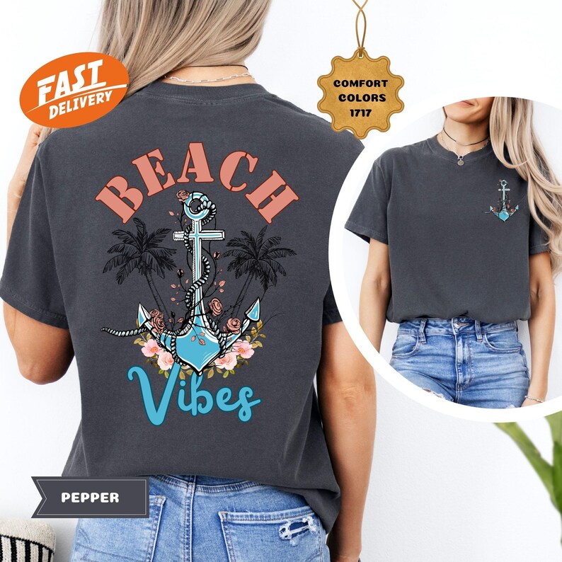 K&ouml;nnte beinhalten: Dunkelgraues T-Shirt mit Strandmotiv. Das Design zeigt einen Anker, Palmen und die Worte "Beach Vibes" in Koralle und T&uuml;rkis. Ein kleineres Ankerdesign befindet sich auf der Vorderseite. Das Shirt ist mit "Pepper" und "Comfort Colors 1717" gekennzeichnet.