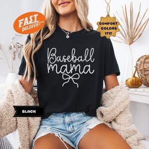 May include: Black t-shirt with the text "Baseball Mama" in a white, cursive font with a bow. The shirt is part of the Comfort Colors 1717 collection. The shirt is paired with denim shorts.