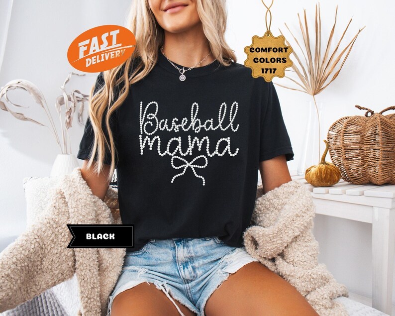 May include: Black t-shirt with the text "Baseball Mama" in a white, cursive font with a bow. The shirt is part of the Comfort Colors 1717 collection. The shirt is paired with denim shorts.