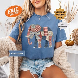 May include: Blue jean t-shirt featuring a patchwork elephant design with a large bow. The elephant is made of various fabrics in red, white, and blue. The shirt has a round neckline and short sleeves. The words "BLUE JEAN" are printed on a blue banner.