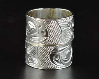 May include: A silver ring with a carved design of two salmon. The ring is wide and has a textured surface.