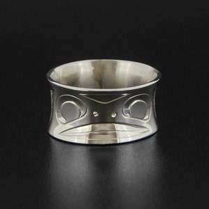 May include: A silver ring featuring a carved design of a stylised face. The ring is wide and has a rounded shape.