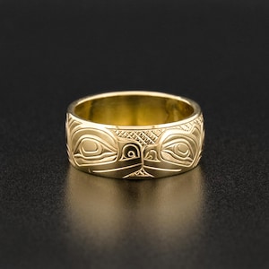 May include: A gold ring with a detailed design of two stylized animal faces. The ring is wide and has a smooth, polished finish.