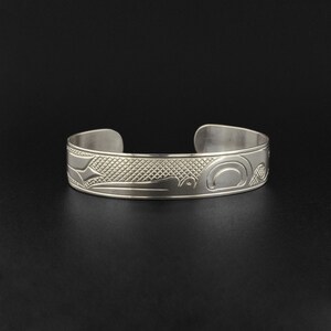 May include: A silver bracelet with a detailed design of a stylized bird or animal. The design is engraved into the metal and has a textured background.