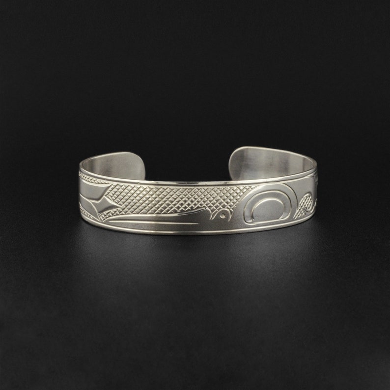 May include: A silver bracelet with a detailed design of a stylized bird or animal. The design is engraved into the metal and has a textured background.