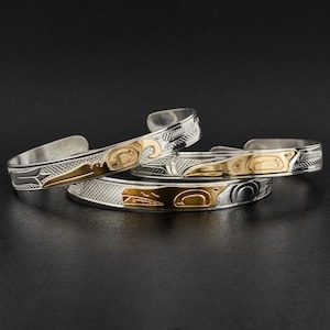 Puede incluir: Tres pulseras de plata con diseños intrincados, que presentan un motivo de pájaro dorado. Las pulseras están apiladas una encima de la otra, mostrando la artesanía detallada.